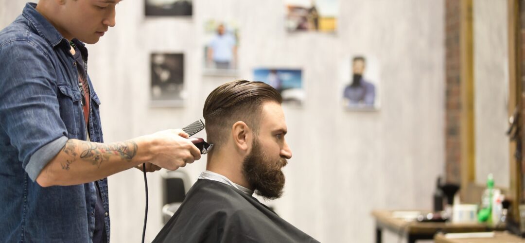 Client in barbershop Side view portrait of handsome young bearded caucasian man getting trendy haircut in modern barbershop. Cool men hair stylist with tattoo "born barber" serving client. Indoors shot